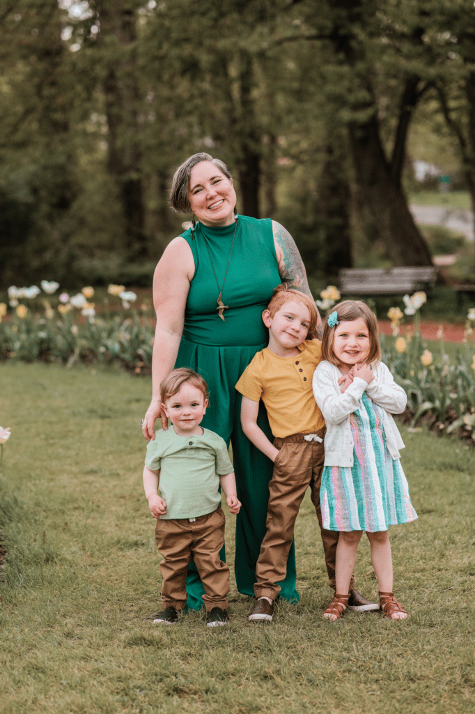 Untitled design Image shows Marlie Schmidt, Physician Assistant, smiling and posing in a grassy area with her 3 young children smiling while standing in front of her.